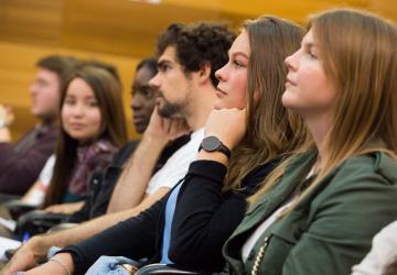 A group of diverse young adults attentively listening in a seminar or lecture setting, seated in a row with a focus on their engaged expressions.
