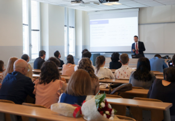 A speaker presents in front of an audience seated at wooden desks in a classroom, with a projector screen displaying a presentation. A bouquet of flowers sits on the desk in the foreground.